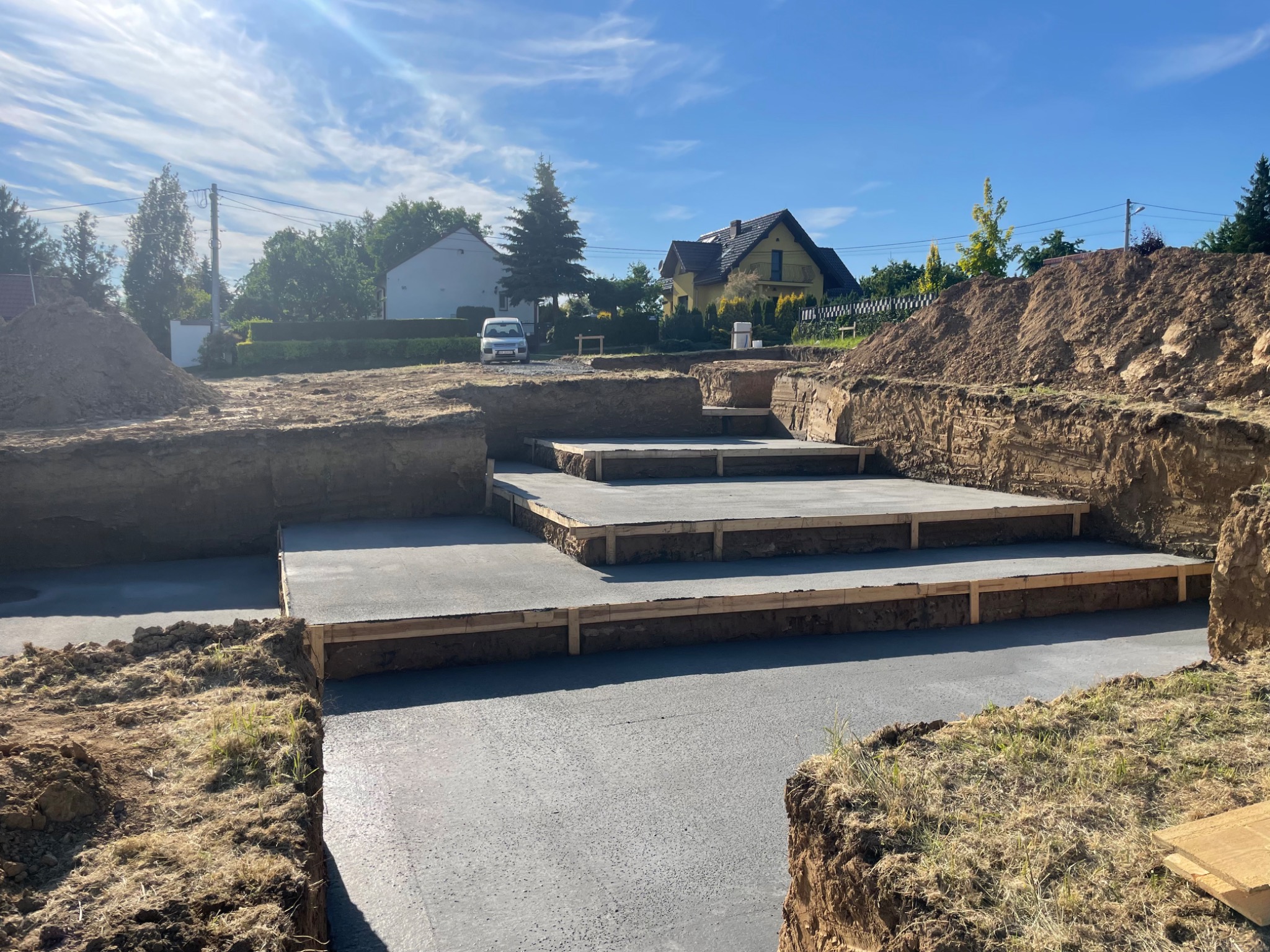 Wylany betonowy fundament z drewnianym szalunkiem na nierównym terenie z widokiem na domy w tle.