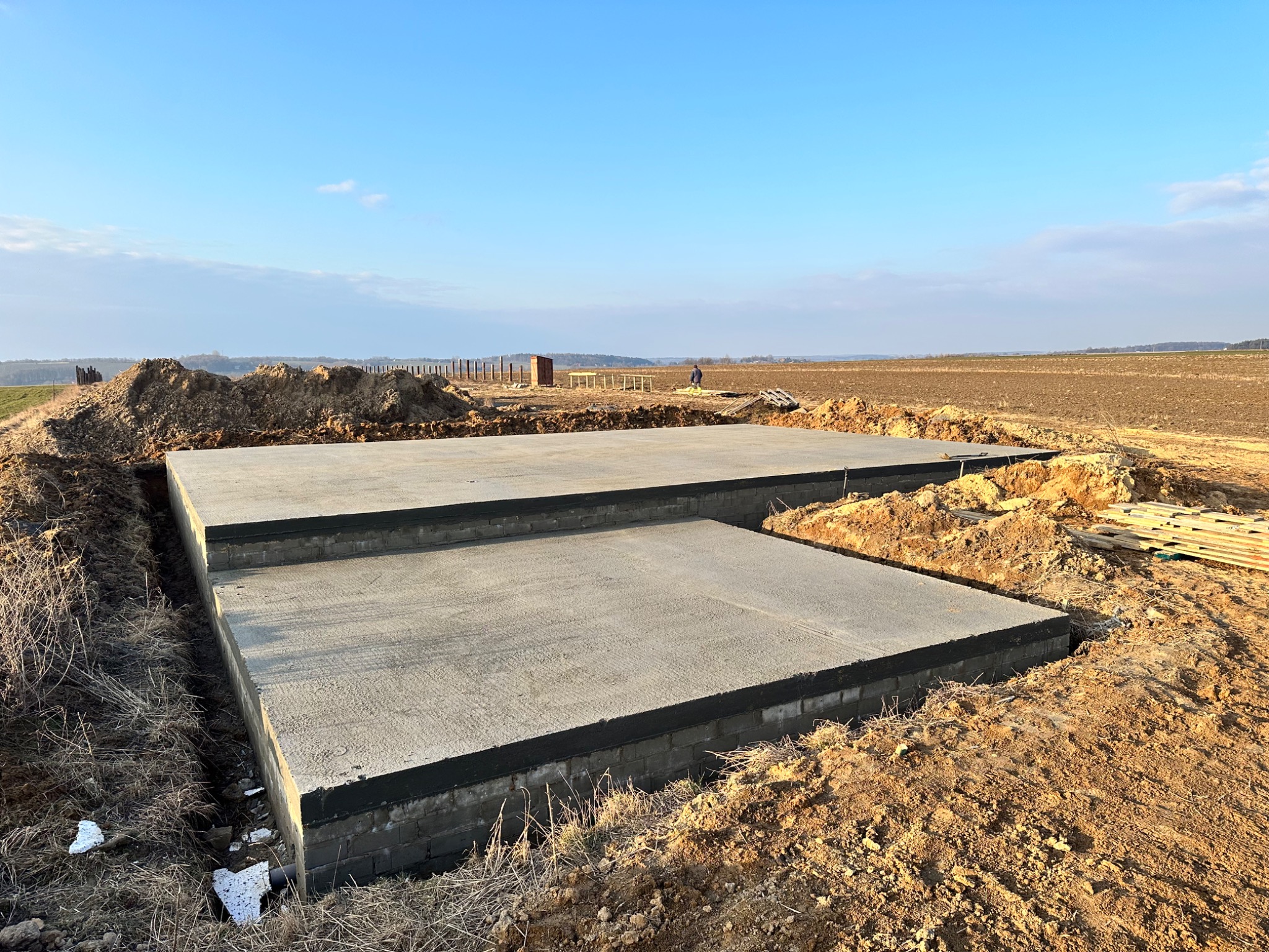 Dwa betonowe fundamenty w wykopie, zabezpieczone czarną izolacją, na tle zaoranego pola i błękitnego nieba z pojedynczym pracownikiem w oddali.