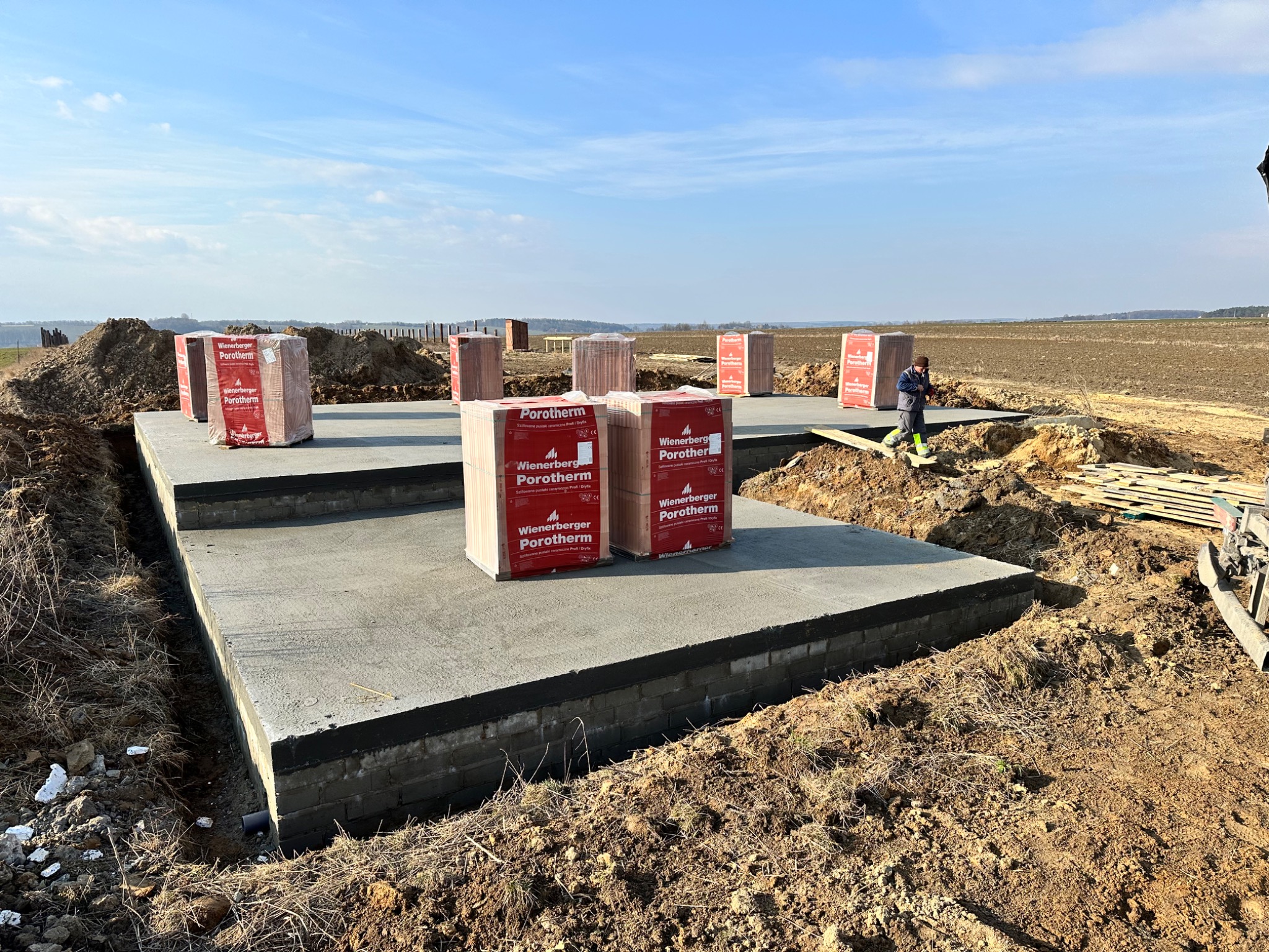 Wylany fundament z bloczków betonowych, zabezpieczony czarną izolacją, z ustawionymi paletami cegieł Porotherm, na rozkopanym terenie budowy, widoczna osoba w oddali.