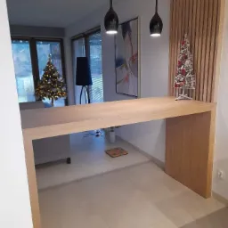 Nowoczesna aranżacja wnętrza z drewnianym blatem przechodzącym w ściankę działową, wiszące czarne lampy, dekoracyjna ścianka z drewnianych listew i choinka na blacie, w tle widoczna choinka...