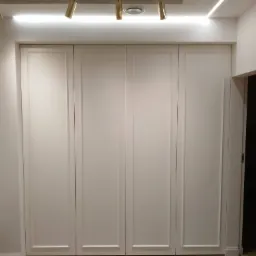 Czterodrzwiowa szafa z białymi frontami, zdobiona delikatnymi frezowaniami, oświetlona punktowymi lampami sufitowymi i listwą LED, na podłodze parkiet w jodełkę.