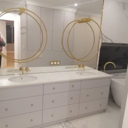 Meblo Łukasz - Biała szafka łazienkowa z dwoma umywalkami, złotymi bateriami i okrągłymi wieszakami na ręczniki, odbicie lustrzane z widoczną osobą trzymającą telefon.