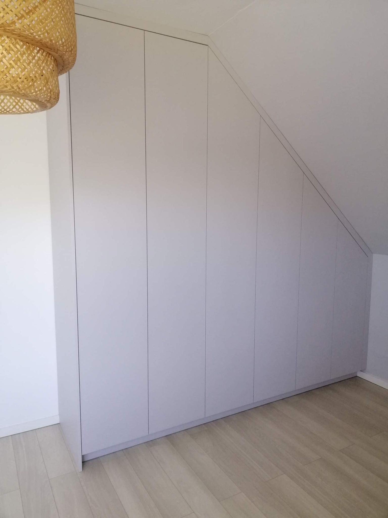 Szara zabudowa szafy z frontami bez uchwytów, dopasowana do skosu poddasza, z widoczną częścią drewnianej podłogi i lampą z wikliny.