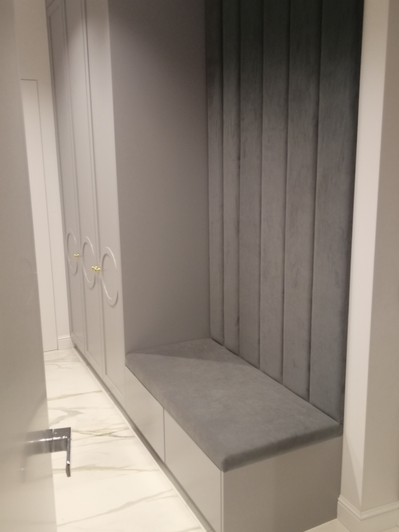 Szara zabudowa w przedpokoju z siedziskiem z tapicerowanym panelem ściennym i szafą z owalnymi, złotymi uchwytami.