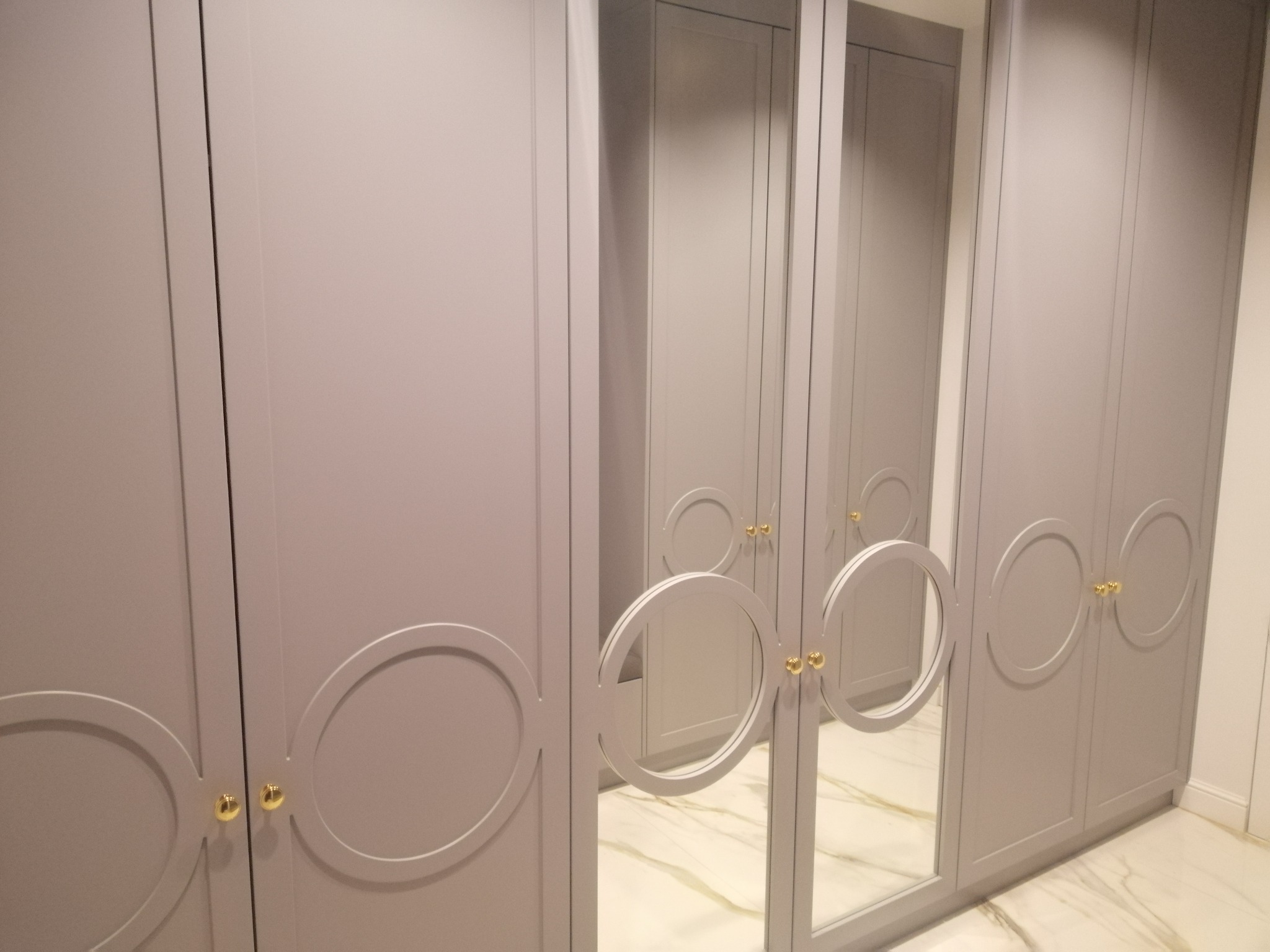 Pojemna, szara szafa z lustrzanymi drzwiami i okrągłymi, złotymi uchwytami, stojąca na marmurowej podłodze.