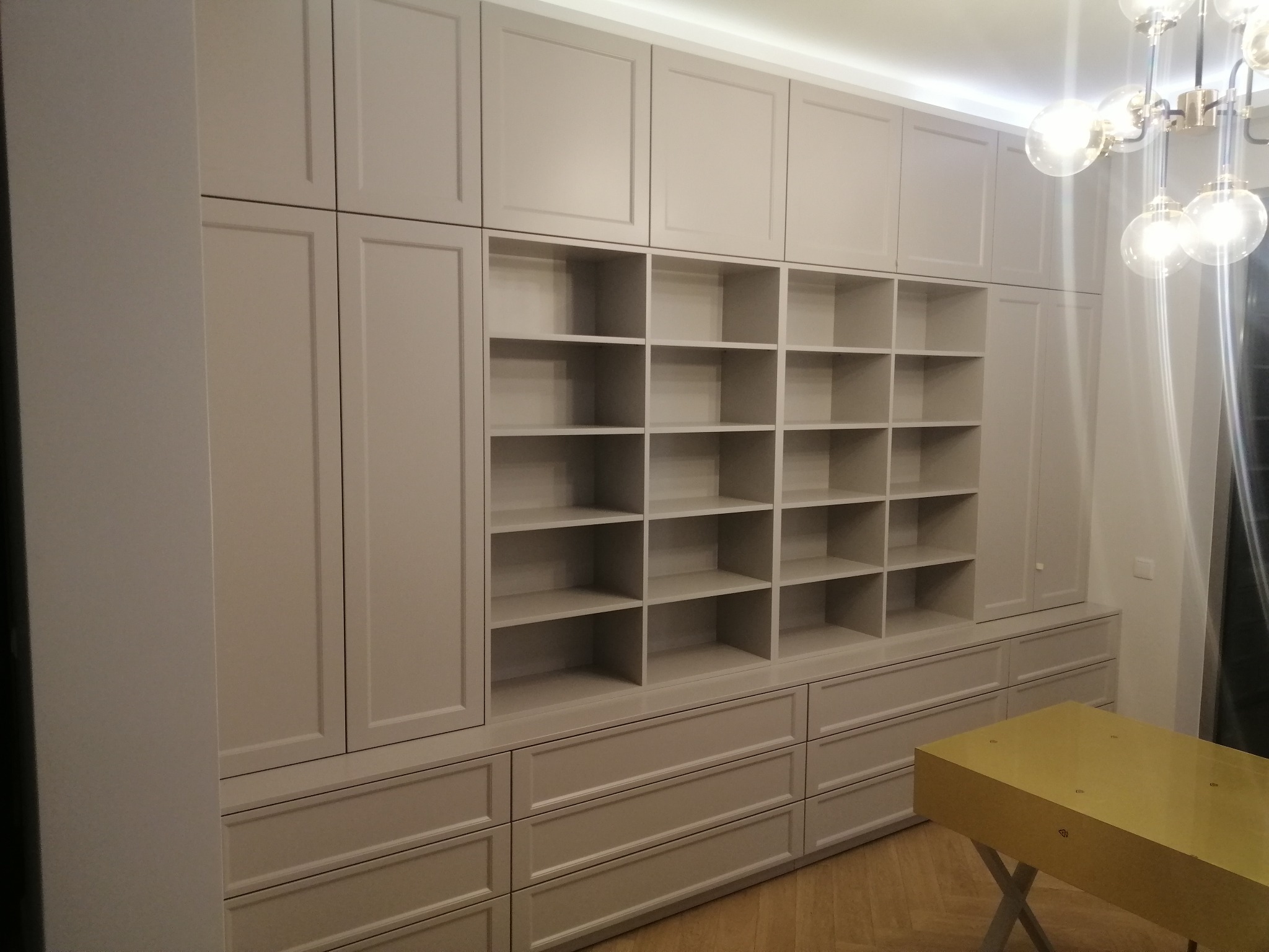 Duża, jasnoszara zabudowa meblowa z licznymi szafkami, szufladami i otwartymi półkami w nowoczesnym wnętrzu z drewnianą podłogą w jodełkę i designerską lampą.