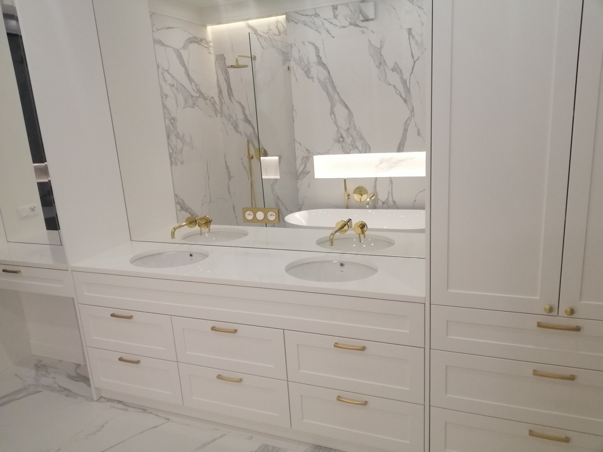 Biała łazienka z umywalkami wpuszczonymi w blat, złotą armaturą i marmurowym tłem w lustrze.