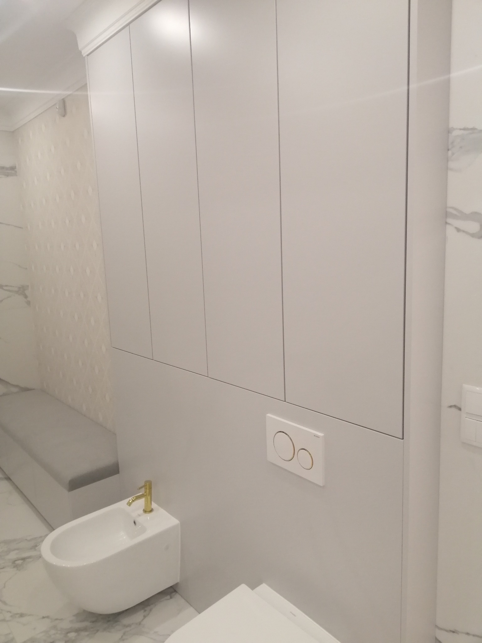 Nowoczesna łazienka z białą zabudową meblową na całej ścianie, wiszącym bidetem z złotą armaturą i marmurową posadzką.