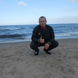 Mężczyzna w sportowym stroju kuca na plaży, pokazując kciuk w górę na tle morza i nieba.