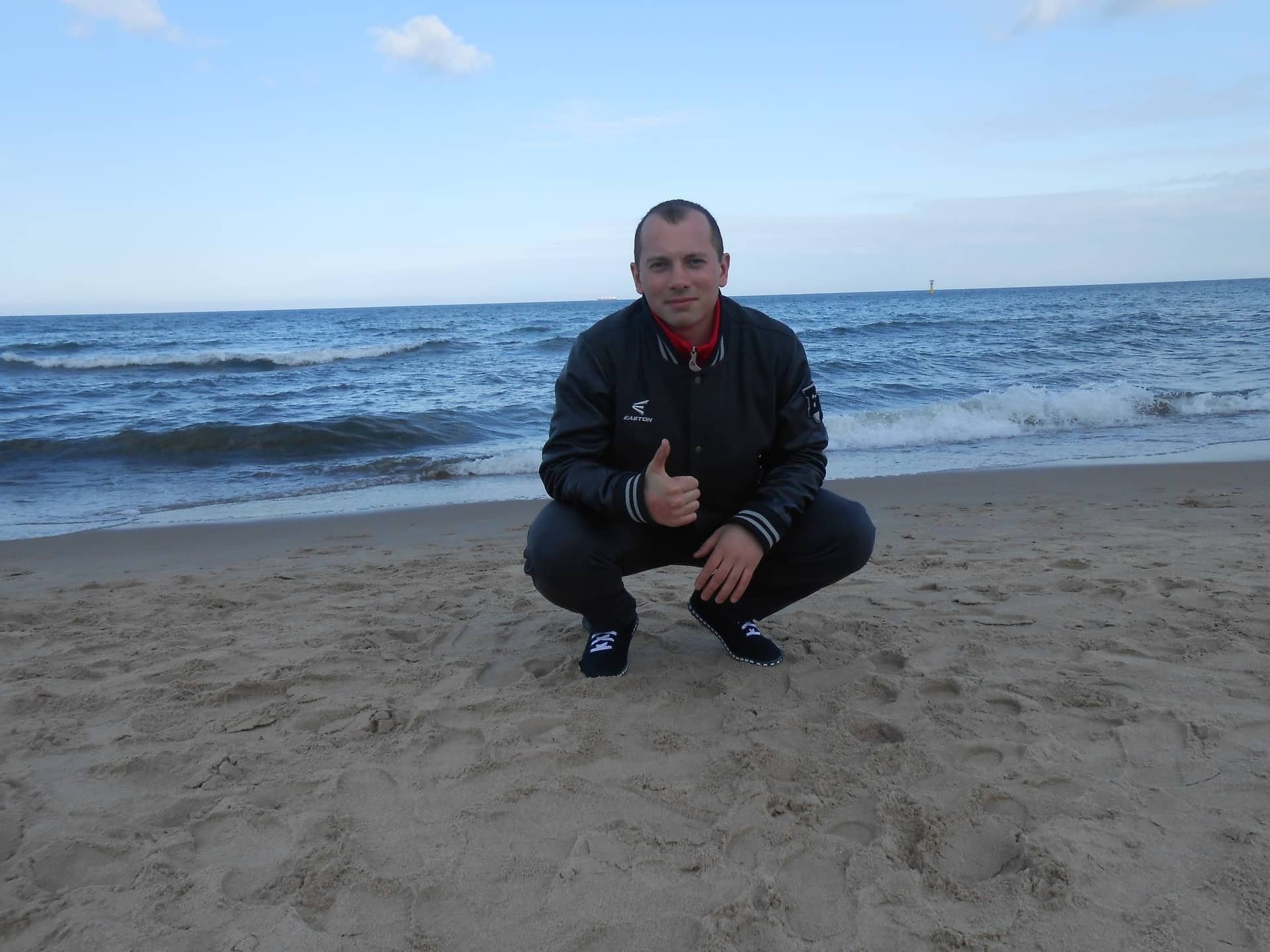 Mężczyzna w sportowym stroju kuca na plaży, pokazując kciuk w górę na tle morza i nieba.