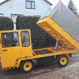 Żółty pojazd transportowy z podnoszoną skrzynią ładunkową z szarą plandeką, widoczny na tle posesji z tujami i budynkiem mieszkalnym.