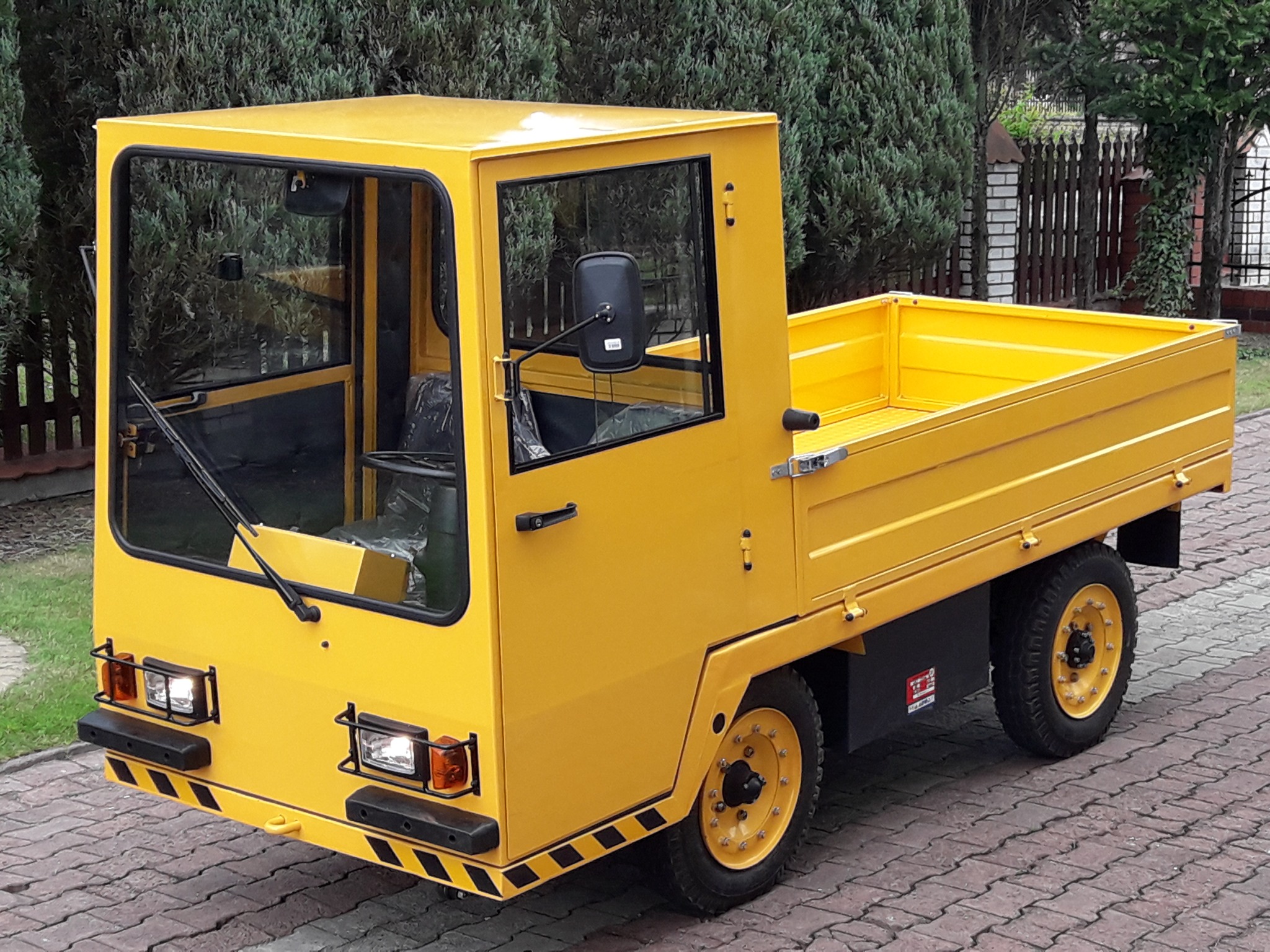 Żółty pojazd transportowy z kabiną i skrzynią ładunkową, widoczny na brukowanej powierzchni, z zielonymi krzewami w tle.
