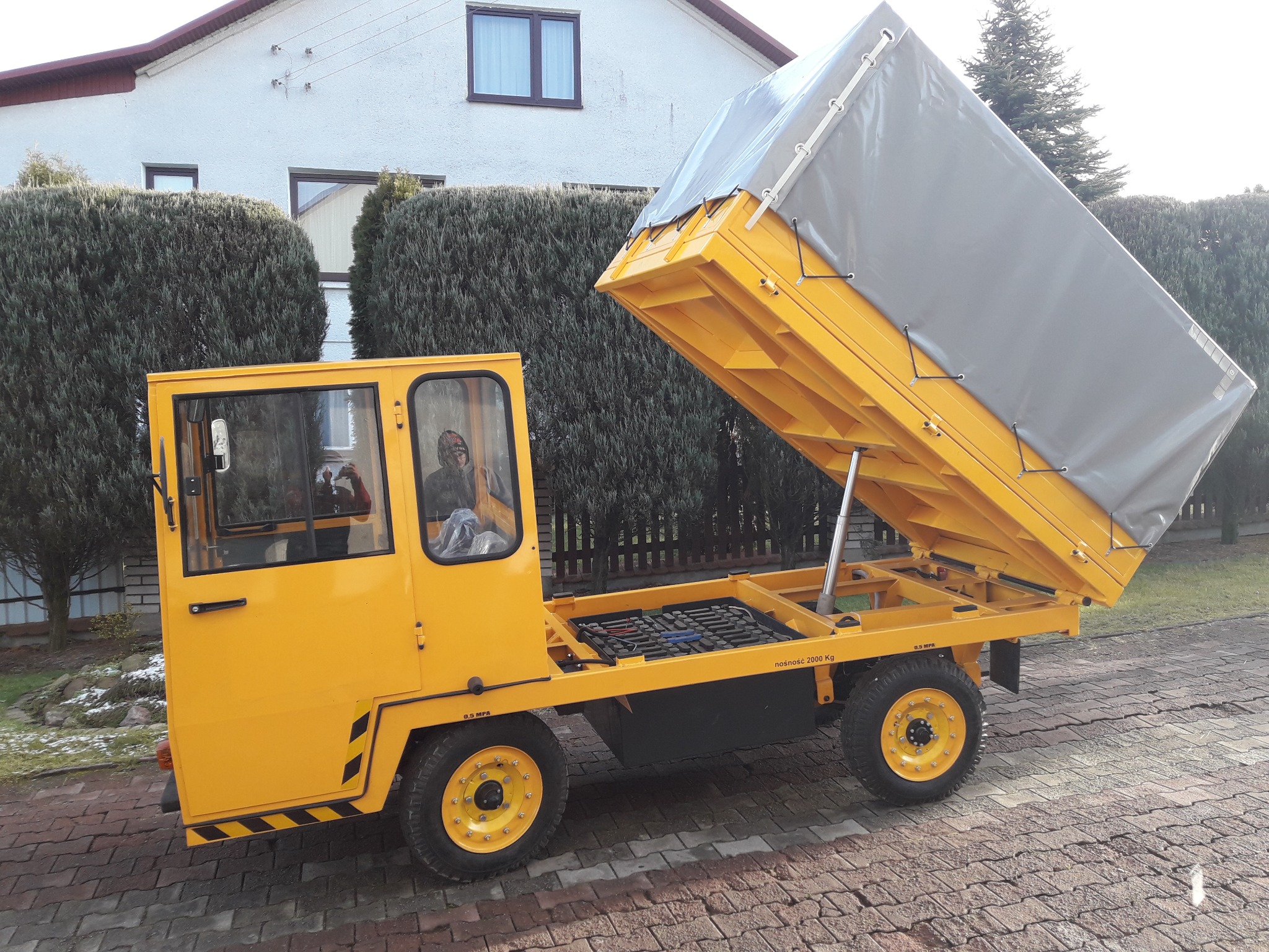 Żółty pojazd transportowy z podnoszoną skrzynią ładunkową z szarą plandeką, widoczny na tle posesji z tujami i budynkiem mieszkalnym.