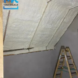 Poddasze z widoczną izolacją natryskową z pianki poliuretanowej, drewniana drabina w rogu pomieszczenia, logo firmy termoizolacyjnej w lewym górnym rogu.