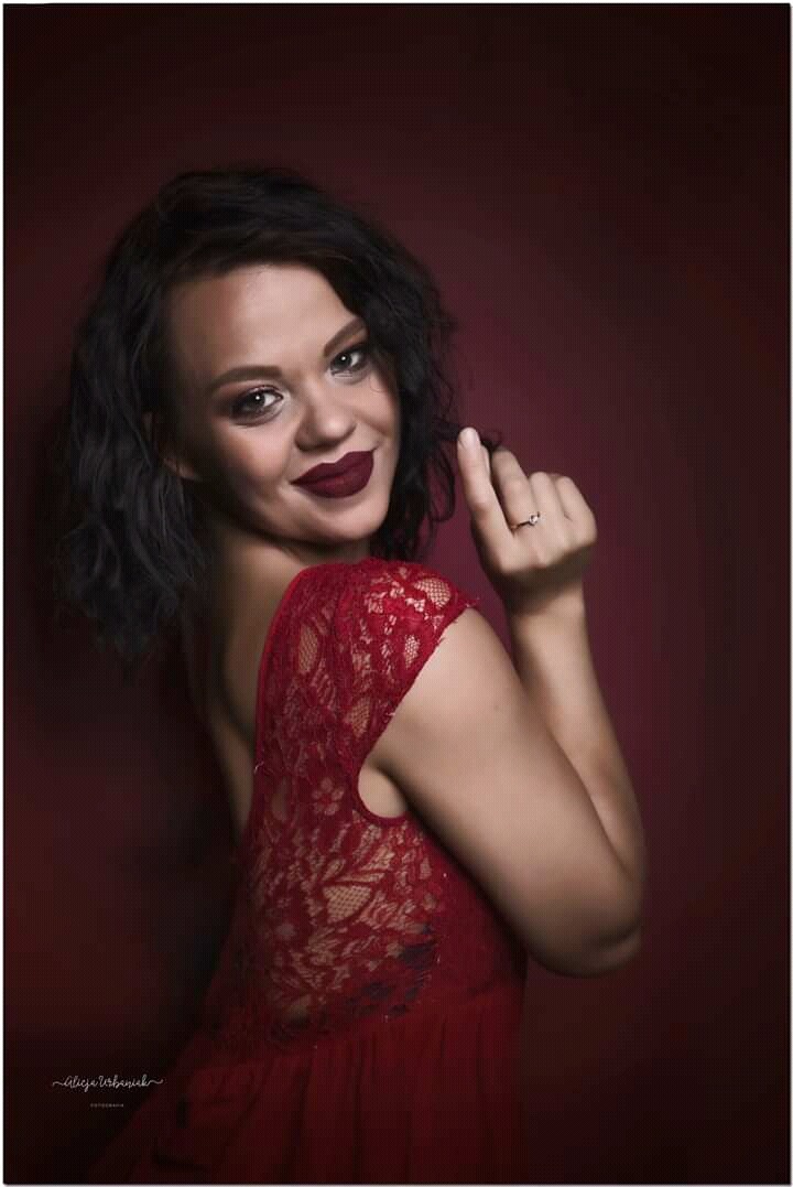 Portret kobiety w czerwonej koronkowej sukience, z ciemnymi włosami i wyrazistym makijażem, pozującej na tle bordowej ściany.