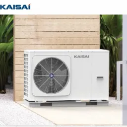 Biała jednostka zewnętrzna urządzenia grzewczego marki Kaisai na metalowej podstawie, umieszczona na tle drewnianej ściany i zielonych roślin. Widoczny fragment białej ściany budynku z drzwiami.