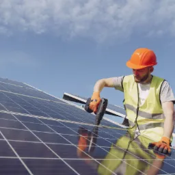 Pracownik w kasku i kamizelce odblaskowej montuje panele słoneczne na dachu, używając wkrętarki. Widoczne niebieskie niebo w tle.