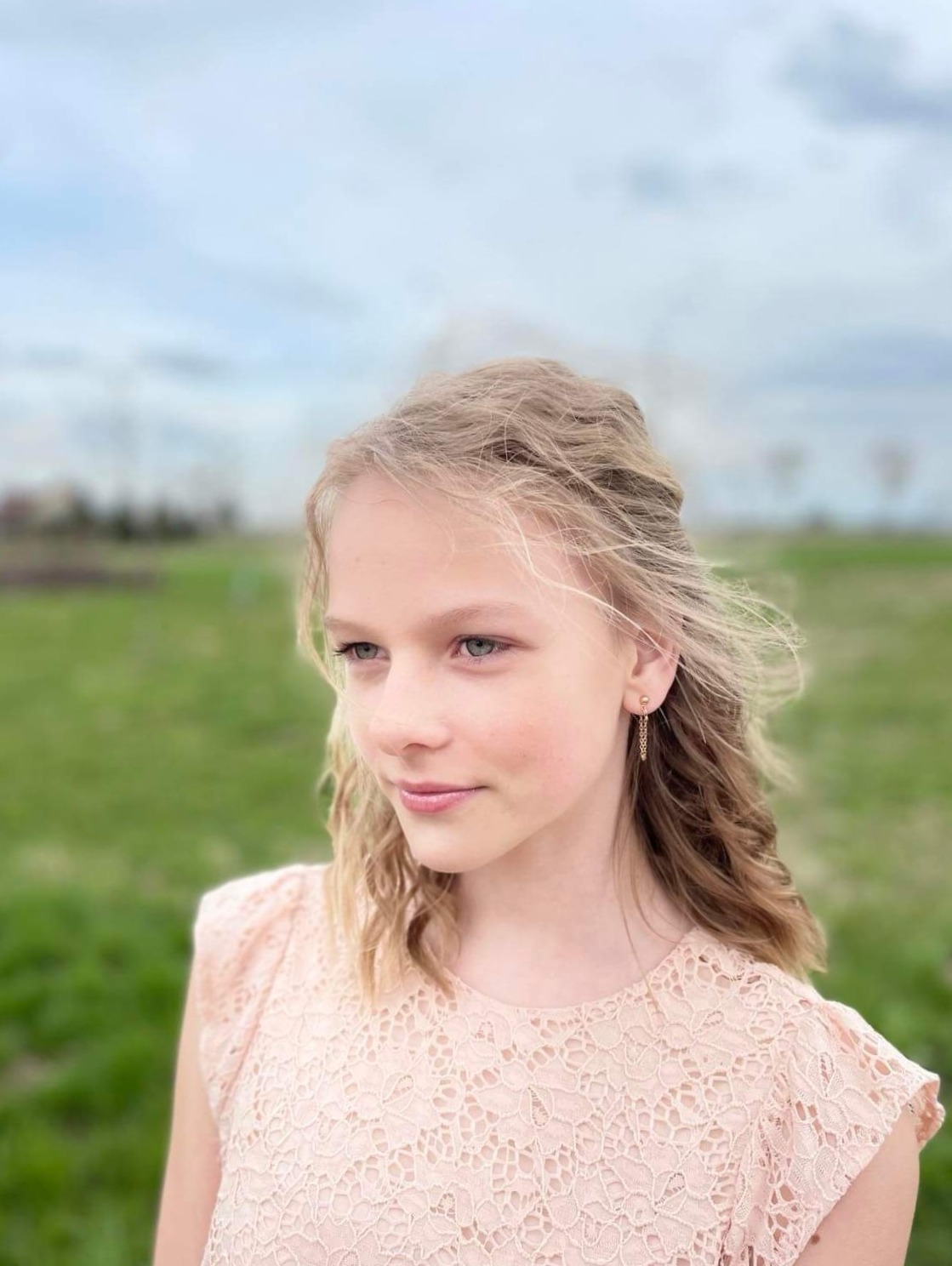 Portret młodej dziewczyny z blond włosami i delikatną złotą kolczykiem łańcuszkową, ubrana w koronkową sukienkę w pastelowym różu, na tle rozmytej zielonej łąki i pochmurnego nieba.