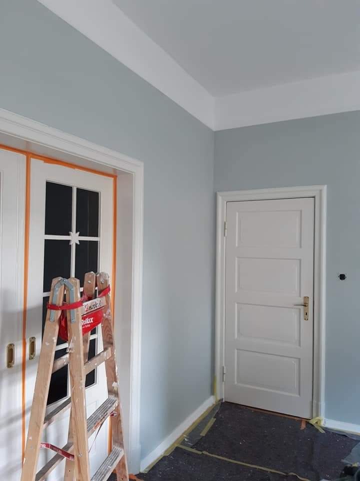 Świeżo pomalowane na szaro ściany pokoju z białymi drzwiami i listwami przypodłogowymi, zabezpieczone folią podłogi, drewniana drabina oparta o przeszklone dwuskrzydłowe drzwi z pomarańczową taśmą...