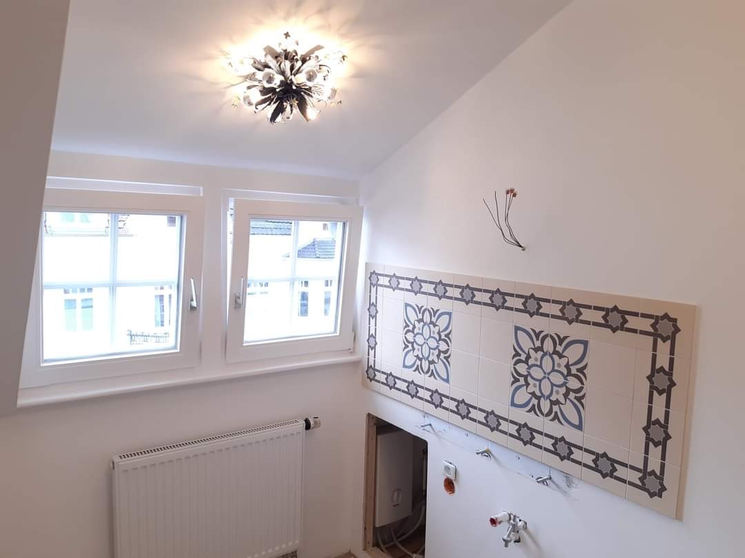 Ściana z dekoracyjnymi płytkami ceramicznymi w trakcie montażu, widoczne wystające kable, grzejnik i okna w jasnym pomieszczeniu.