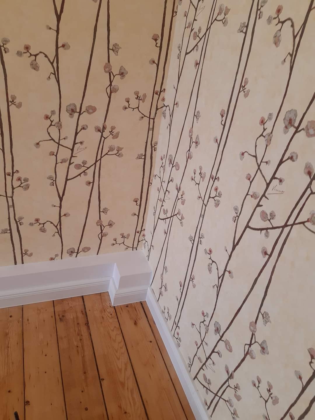 Wnętrze pokoju z drewnianą podłogą, białymi listwami przypodłogowymi i ścianami pokrytymi tapetą w jasnym odcieniu z motywem brązowych gałązek i drobnych, różowych kwiatów.