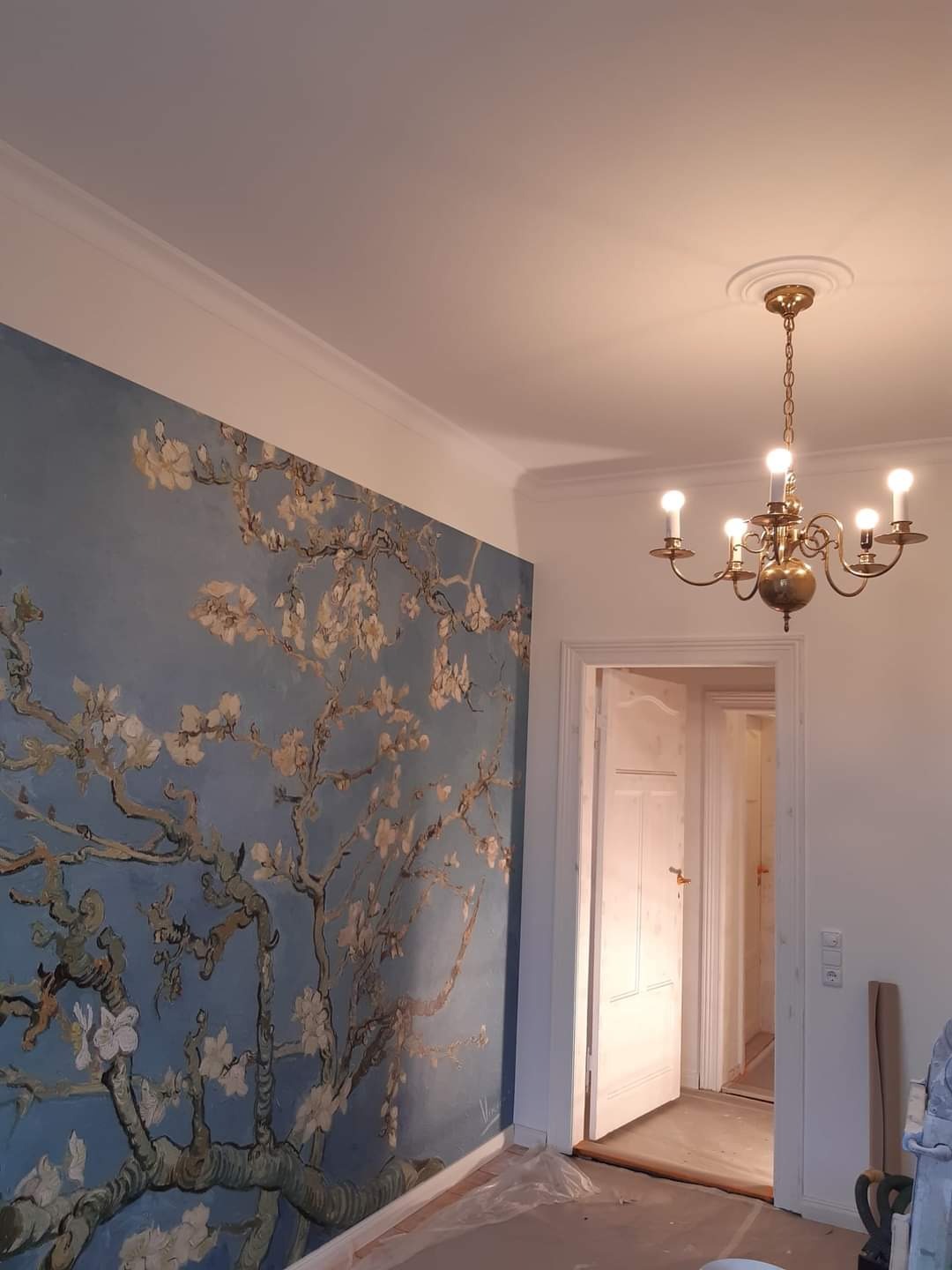 Ściana w pokoju z reprodukcją obrazu kwitnącego migdałowca Vincenta van Gogha, ozdobny żyrandol w stylu retro, widoczne zabezpieczenia podłogi folią malarską.