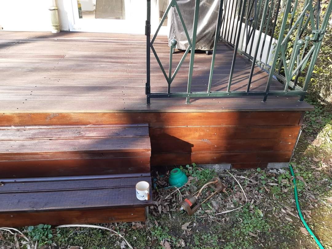 Drewniany taras z metalową balustradą, widoczne schody wejściowe, kubek na schodach, zraszacze ogrodowe i wąż obok tarasu.