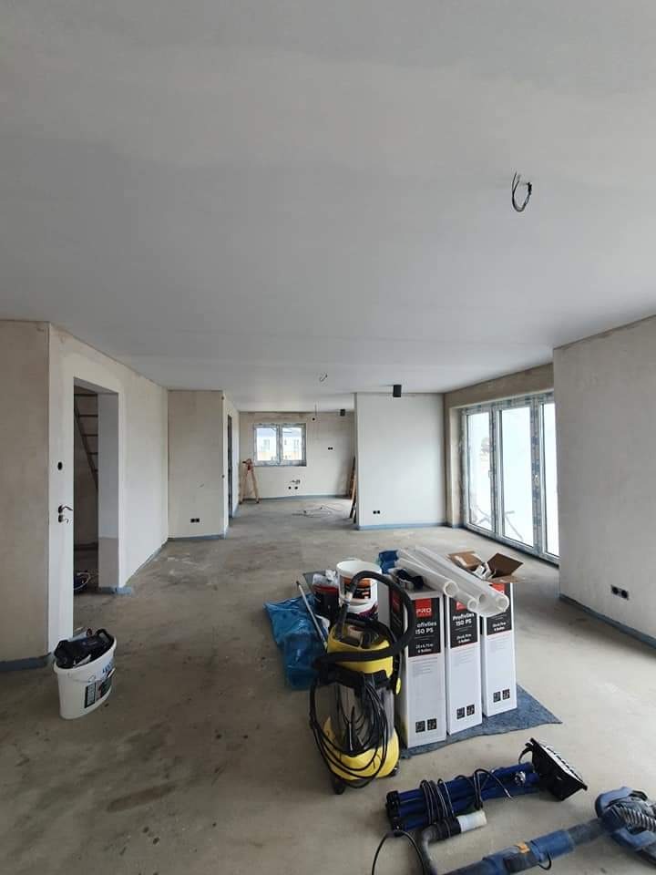 Wnętrze mieszkania w trakcie prac wykończeniowych: surowe ściany przygotowane do malowania, widoczne narzędzia, odkurzacz przemysłowy, puszki z farbą i listwy PCV na podłodze, duże okna balkonowe.