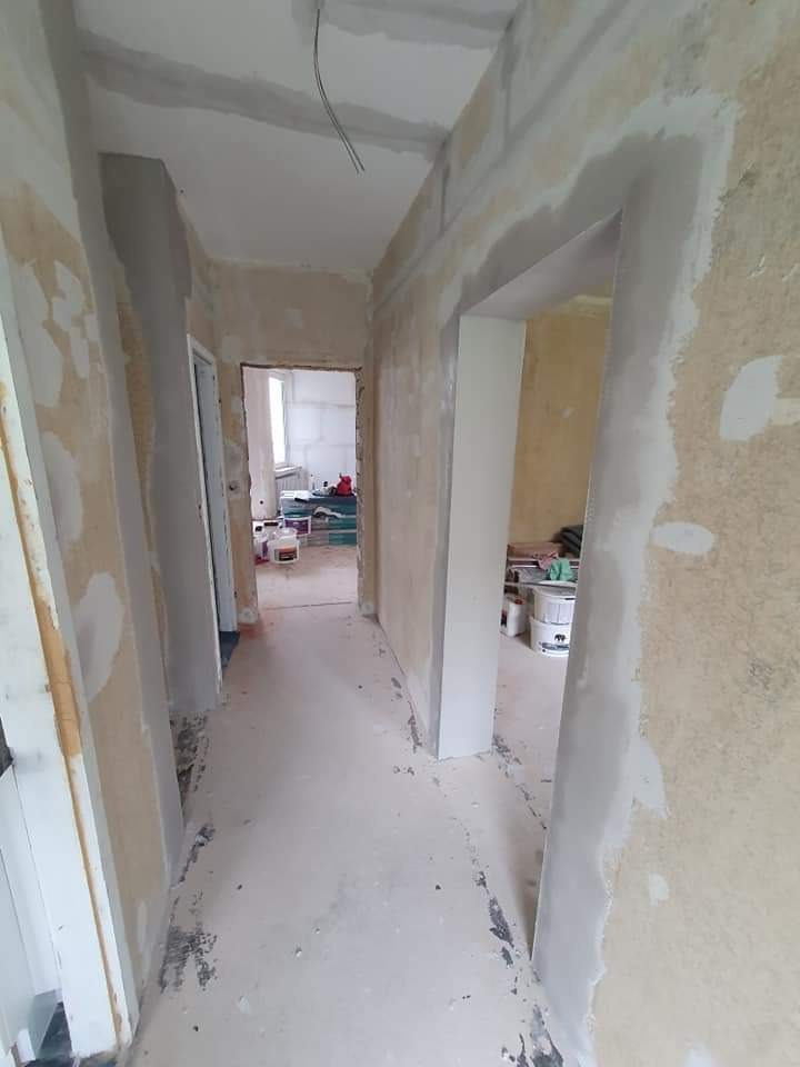 Korytarz w trakcie przygotowania ścian do malowania, widoczne ślady po szpachlowaniu i gładzi, odsłonięte kable na suficie, w tle pomieszczenie z wiadrami i narzędziami.