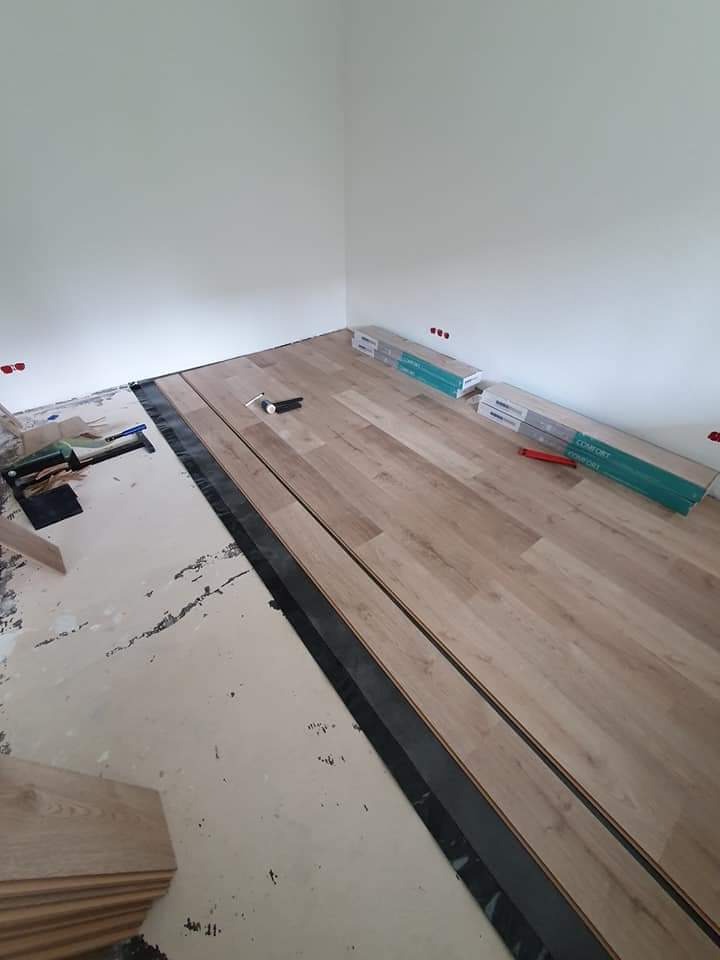 Układanie paneli podłogowych w trakcie pracy: widoczne panele Comfort, czarna folia izolacyjna, narzędzia i częściowo wykończona podłoga w rogu pomieszczenia z białymi ścianami.