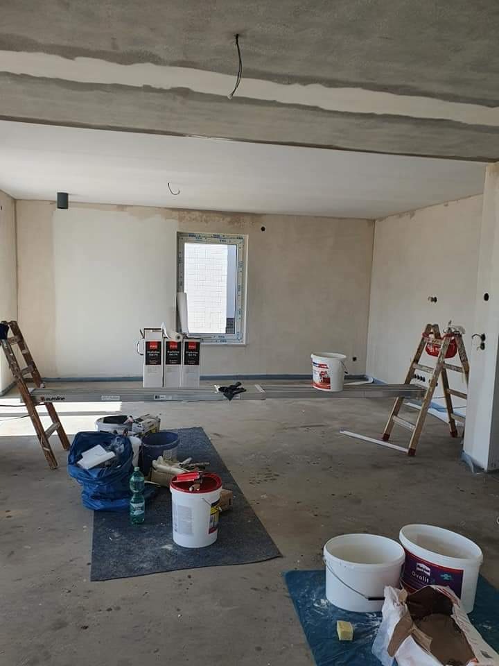 Surowy stan wnętrza w trakcie prac wykończeniowych: ściany częściowo pokryte gładzią, widoczne drabiny, wiadra z farbą i narzędzia malarskie na podłodze zabezpieczonej folią malarską, okno...