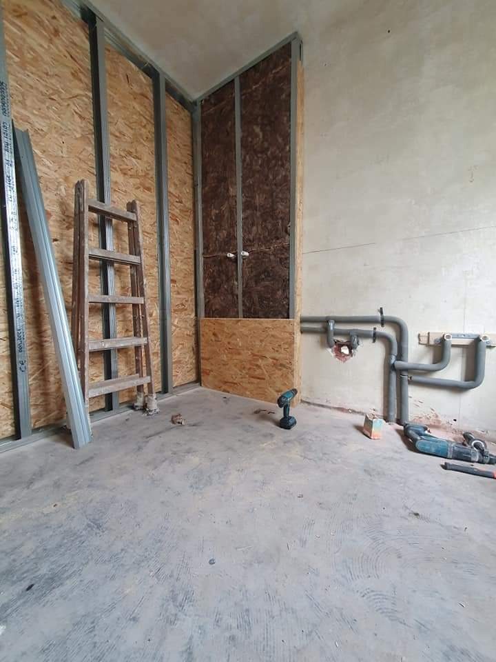 Wnętrze łazienki w trakcie prac przygotowawczych, widoczne płyty OSB na ścianach, metalowa konstrukcja szkieletowa, drabina, instalacja wodna, wiertarka akumulatorowa i inne narzędzia na surowym...