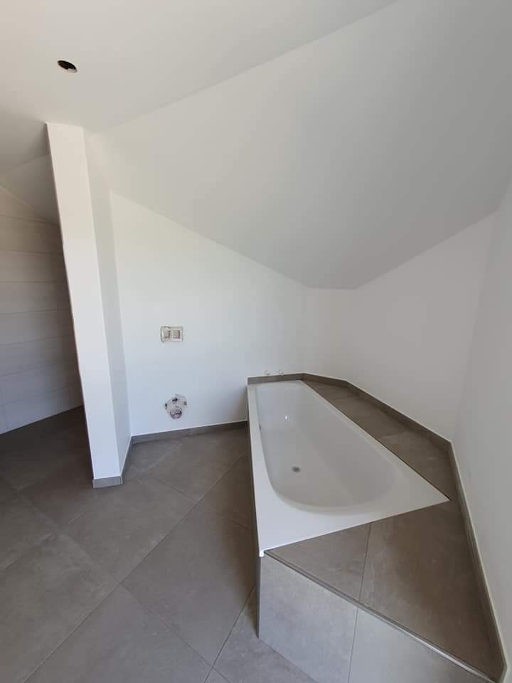 Nowoczesna łazienka w trakcie prac wykończeniowych: biała wanna narożna obudowana szarymi płytkami, szara podłoga z płytek, białe ściany z widocznymi puszkami instalacyjnymi, sufit ze skośnym...