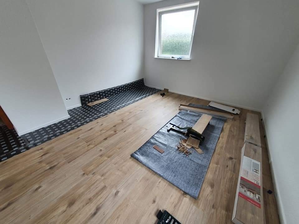 Układanie paneli podłogowych w jasnym pokoju z oknem, widoczna mata podkładowa, narzędzia i opakowania paneli.