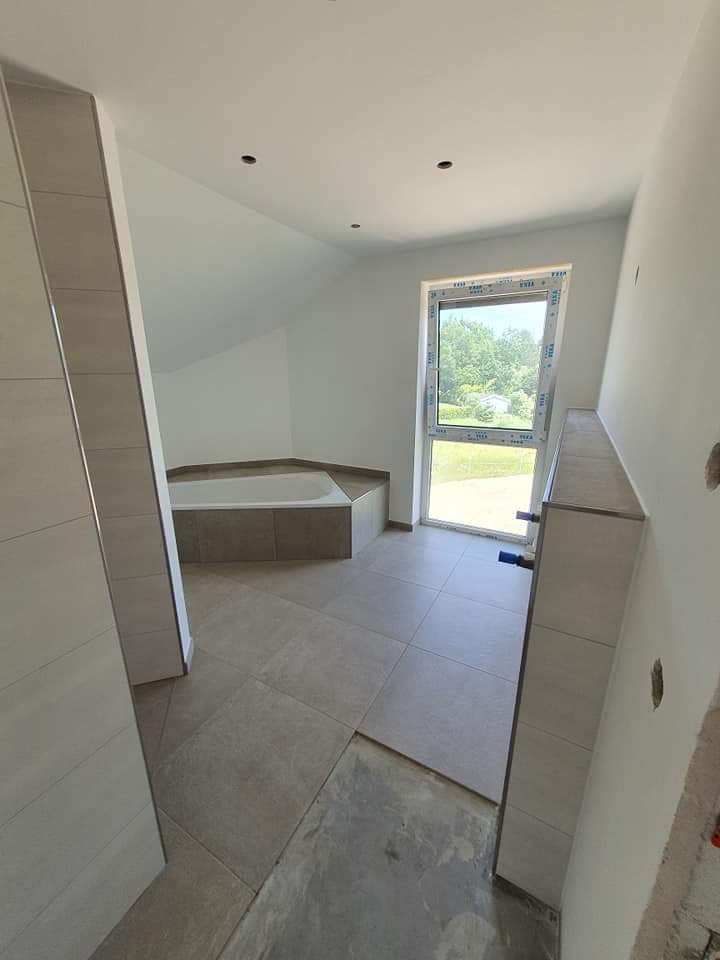 Nowoczesna łazienka w trakcie wykańczania, z widoczną wanną w zabudowie z płytek, jasnymi płytkami podłogowymi i częściowo wykończonymi ścianami, z oknem na zewnątrz i wystającymi rurami instalacji.