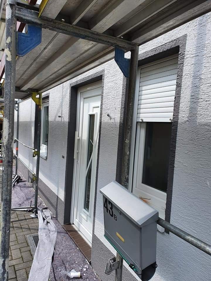 Elewacja budynku z szarym tynkiem strukturalnym, białą stolarką okienną i drzwiową, skrzynka pocztowa z numerem 43B, rusztowanie w trakcie prac.