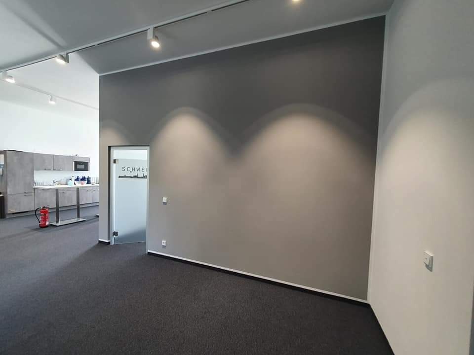 Wnętrze biura po malowaniu ścian na szaro, z widocznym oświetleniem punktowym, szarą wykładziną i częściowo widoczną kuchnią w tle.