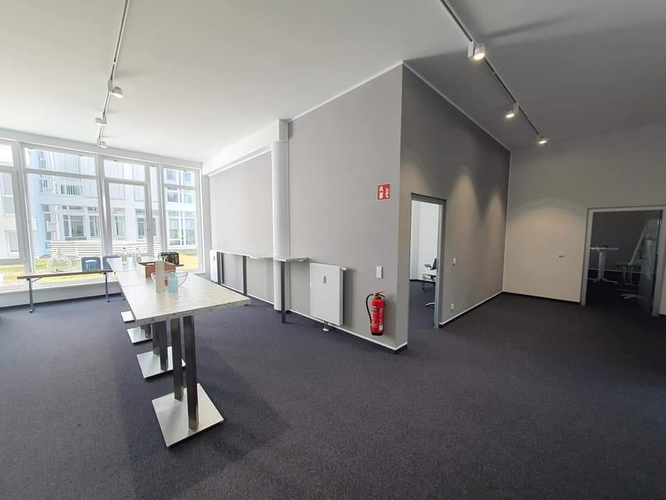 Jasne, przestronne biuro z szarym dywanem, wysokimi stołami i dużymi oknami, widoczne grzejniki, gaśnica i drzwi do innych pomieszczeń.