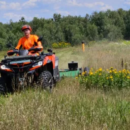 Operator w pomarańczowym kombinezonie i kasku na pomarańczowym quadzie z zamontowaną kosiarką bijakową, w trakcie koszenia wysokiej trawy na łące, widoczne żółte kwiaty i drzewa w tle.