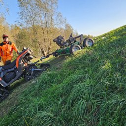 DREWEX S.C. - Operator w pomarańczowym kombinezonie stoi obok pomarańczowo-czarnego quada gąsienicowego, który ciągnie zieloną kosiarkę na zboczu porośniętym wysoką trawą.
