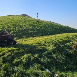 DREWEX S.C. - Pomarańczowy quad z gąsienicami stoi na zboczu zielonego wzgórza, obok leży kosa spalinowa, trawa świeżo skoszona.
