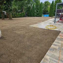 Przygotowany teren pod trawnik, świeżo rozsypana ziemia, widoczne obrzeże z kostki brukowej, w tle tuje i elementy architektury ogrodowej.