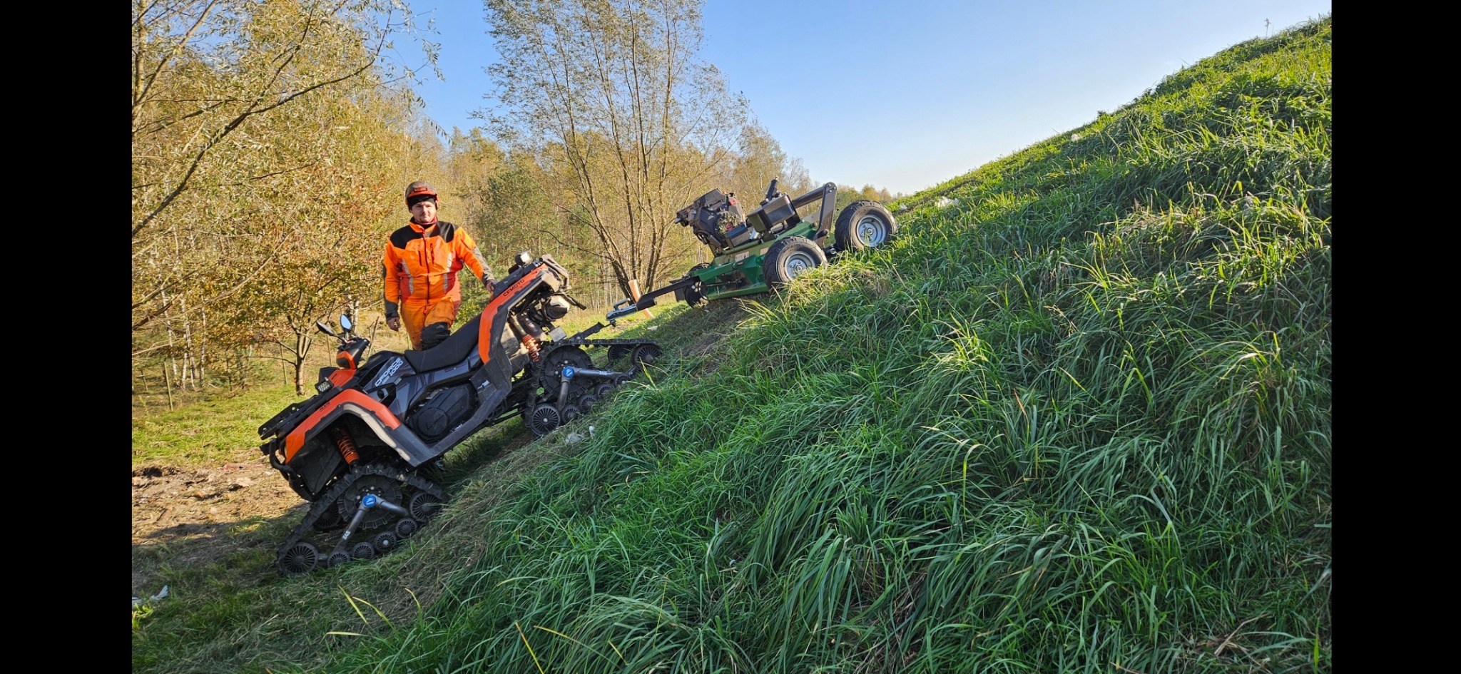 Operator w pomarańczowym kombinezonie stoi obok pomarańczowo-czarnego quada gąsienicowego, który ciągnie zieloną kosiarkę na zboczu porośniętym wysoką trawą.