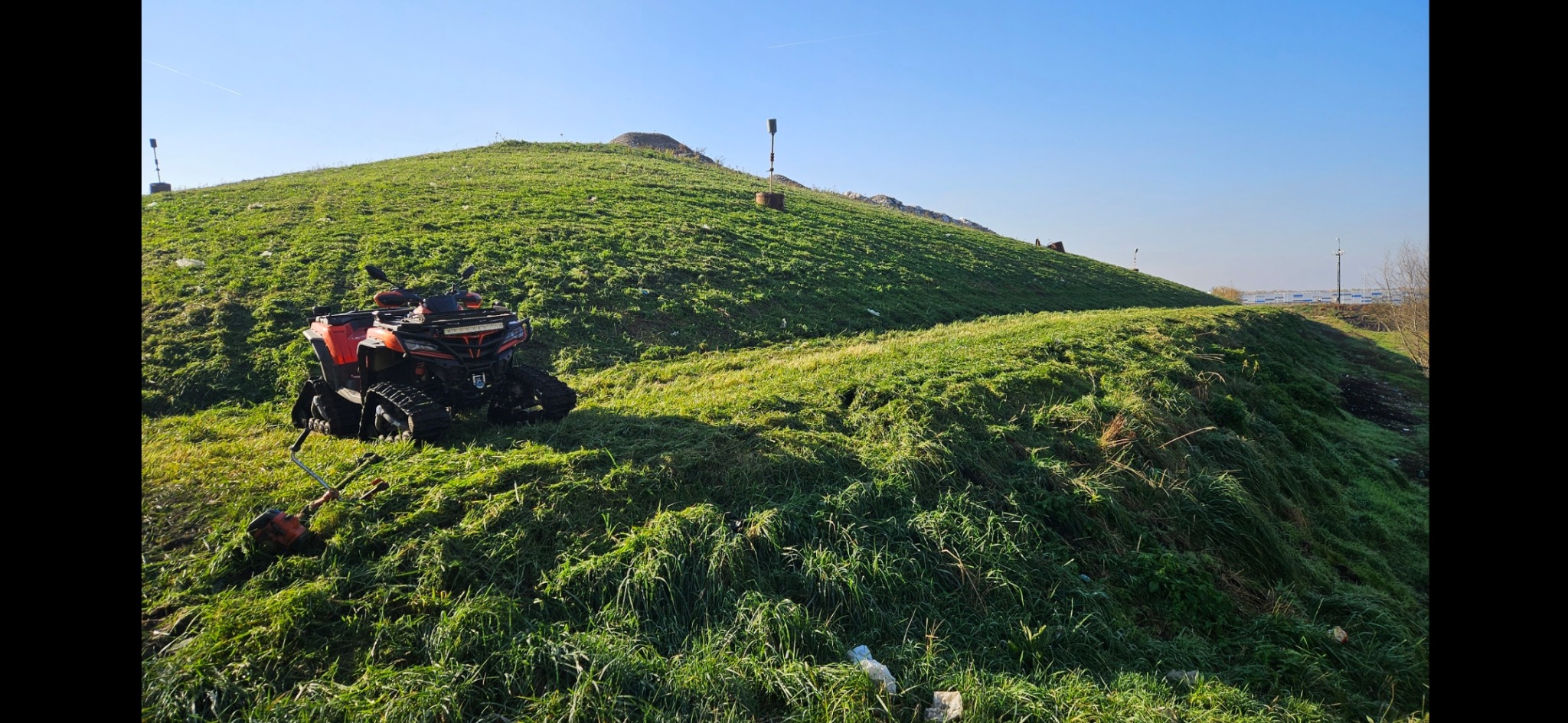 Pomarańczowy quad z gąsienicami stoi na zboczu zielonego wzgórza, obok leży kosa spalinowa, trawa świeżo skoszona.
