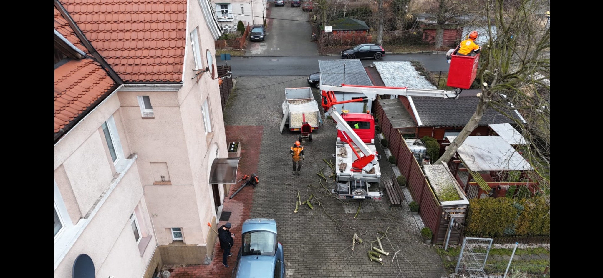 Wycinka drzewa z podnośnika koszowego na posesji, widoczny rębakiem do gałęzi, pracownicy w odzieży ochronnej, widok z góry na teren po wycince.
