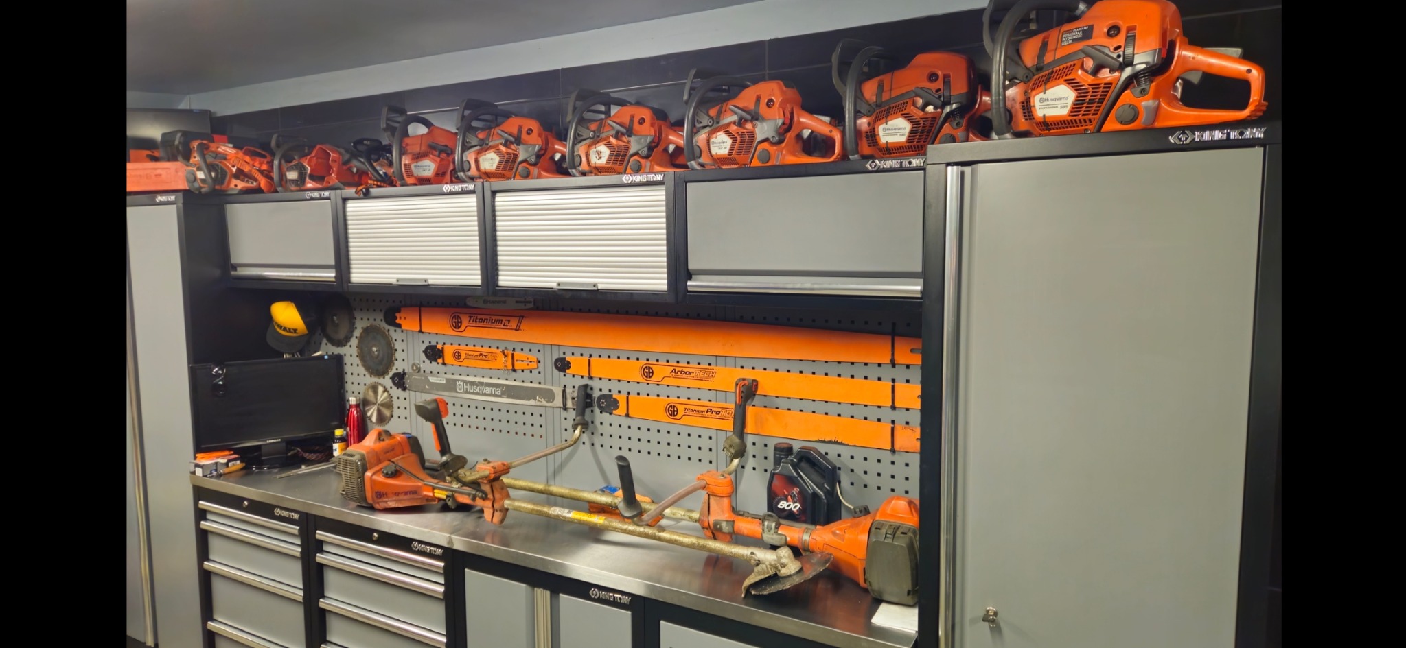 Warsztat z narzędziami ogrodniczymi: rzędy pomarańczowych pilarek łańcuchowych Husqvarna nad szafkami, pod nimi metalowa ściana z zawieszonymi ostrzami, na blacie dwie podkaszarki spalinowe i olej...