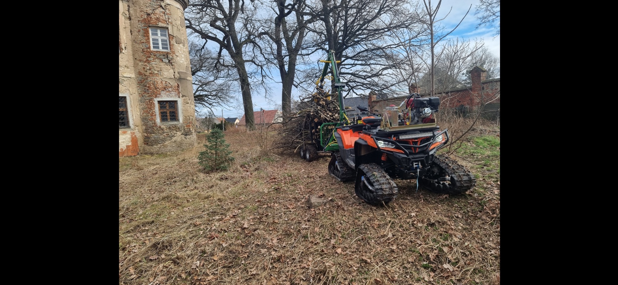 Pomarańczowy pojazd gąsienicowy z przyczepką pełną gałęzi na tle starego budynku w Reńskiej Wsi.