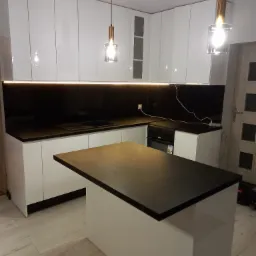 Nowoczesna kuchnia w Radomsku z białymi, połyskującymi frontami i czarnym blatem z kamienia, oświetlona ledami i wiszącymi lampami.