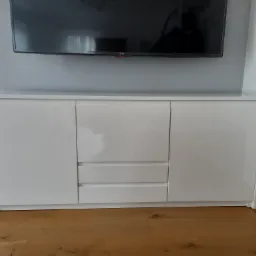 Biała, lakierowana komoda RTV z trzema szufladami i dwojgiem drzwi, stojąca na drewnianej podłodze, pod wiszącym telewizorem LG.