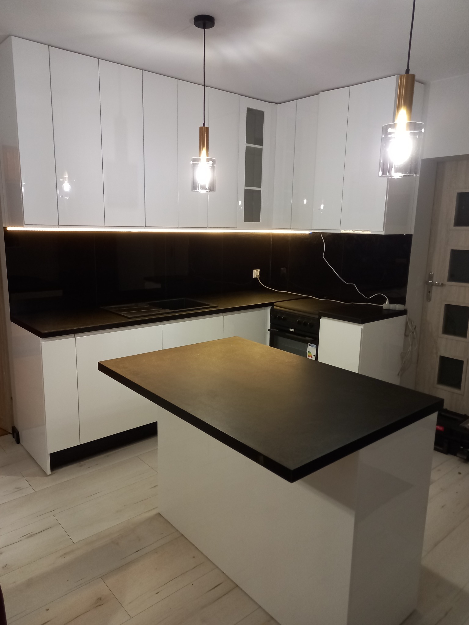Nowoczesna kuchnia w Radomsku z białymi, połyskującymi frontami i czarnym blatem z kamienia, oświetlona ledami i wiszącymi lampami.